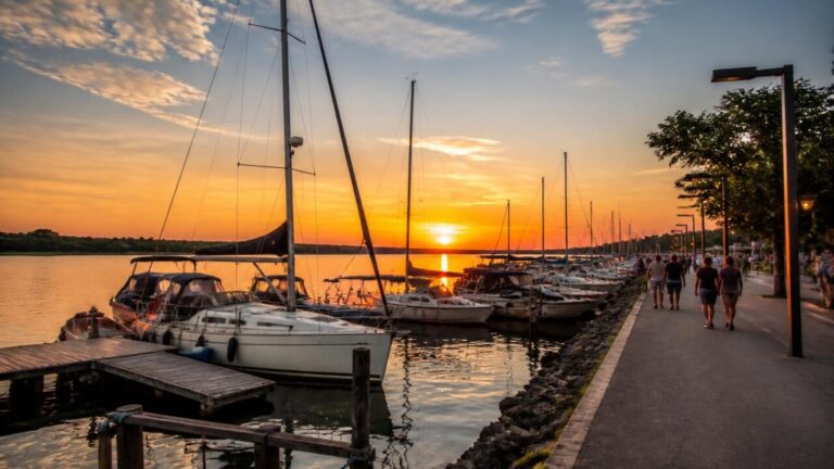 Veleiros e pequenas embarcações atracadas em uma marina ao entardecer, com pessoas na beira observando o movimento.