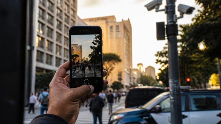 Mão segurando um celular gravando uma rua com câmeras e agentes em atividade policial ao fundo
